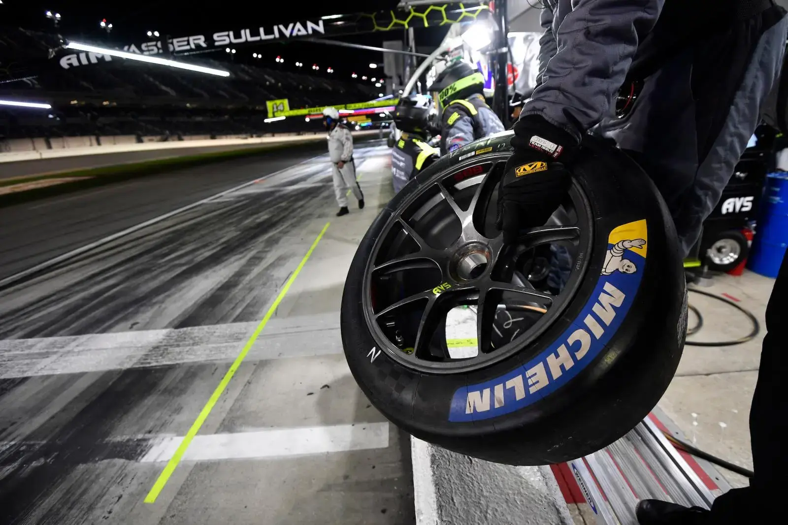 A pit crew member holding a Michelin performance race tire in the pit lane at night.